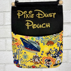 May include: A black fabric hanging pouch with the text Pixie Dust Pouch in gold glitter lettering. The lower pocket features a yellow background with a colorful pattern of a cruise ship, cartoon characters, and fireworks, suspended by a black cord.