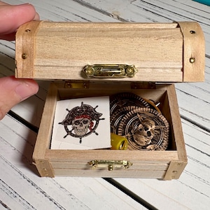 May include: A small, open wooden treasure chest with brass hardware. Inside are pirate-themed items, including coins and a skull and crossbones sticker. The chest is made of light-colored wood.