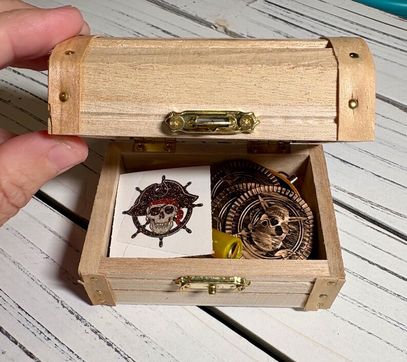 May include: A small, open wooden treasure chest with brass hardware. Inside are pirate-themed items, including coins and a skull and crossbones sticker. The chest is made of light-colored wood.