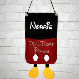 May include: A felt wall hanging with a black top section and a red bottom section, featuring the name "Norris" in white script. The bottom section reads "Pixie Dust Pouch" in gold glitter script, with Mickey Mouse-inspired details.