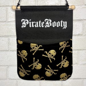 May include: A black fabric wall hanging with a pocket featuring a gold skull and crossbones pattern. The top section displays the white text Pirate Booty in a gothic font. The piece is suspended from a wooden dowel with a black cord for hanging.
