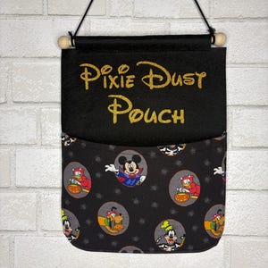 May include: A black fabric wall hanging pouch featuring the gold glitter text Pixie Dust Pouch. The lower pocket section is made of black fabric with a repeating pattern of Disney characters in Halloween costumes, including Mickey Mouse and Pluto.