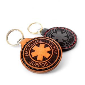 May include: Two leather keychains with a red stitched border. One keychain is brown with the text "Emotional Support Vehicle" and a medical symbol. The other keychain is black with the text "Emotional Support Vehicle" and a medical symbol.