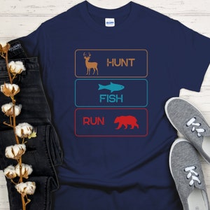 May include: A navy blue t-shirt with a graphic of three buttons. The buttons are labeled "Hunt", "Fish", and "Run". The button for "Hunt" has a deer icon, the button for "Fish" has a fish icon, and the button for "Run" has a bear icon.