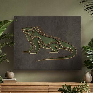 Op de afbeelding: Een groen en goudkleurig leguaan kunstwerk op een donkergrijze vierkant. De leguaan is een gelaagd ontwerp met een gouden omtrek en een groen interieur. Het kunstwerk wordt tentoongesteld boven een houten kast in een kamer met groene muren.