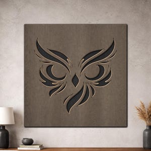 May include: A square wooden wall art piece featuring a stylized owl design. The owl's face is carved out of the wood, with a dark background. The artwork is a neutral brown color, with a detailed, decorative design.