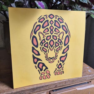 Op de afbeelding: Een vierkante print met een gestileerde grafische illustratie van een jaguar in een voorwaartse houding. Het ontwerp gebruikt een gedurfd kleurenpalet van donkergrijze contouren gevuld met perzikkleurige vormen op een effen, felgele achtergrond.