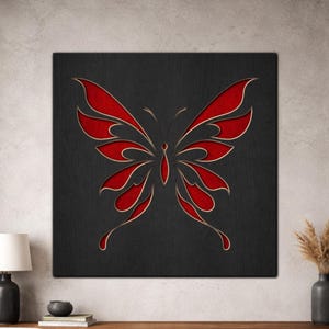 Op de afbeelding: Een vierkant, zwart kunstwerk voor aan de muur met een rood vlinderontwerp met goudkleurige contouren. De vleugels van de vlinder zijn ingewikkeld uitgesneden, waardoor een gelaagd effect ontstaat. Het kunstwerk wordt weergegeven op een getextureerd oppervlak.