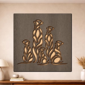 Op de afbeelding: Houten wandkunst met een familie stokstaartjes. De stokstaartjes zijn uitgesneden uit licht hout en geplaatst tegen een donkergrijze achtergrond. Het vierkante kunstwerk heeft een natuurlijke, rustieke uitstraling.