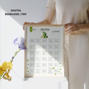 May include: A printable 30-day detox challenge chart with a green juice illustration. The chart is framed in a light wood frame and is being held by a person.