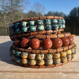 May include: A stack of three leather wrap bracelets with different colored beads. The top bracelet has blue and white beads, the middle bracelet has brown beads, and the bottom bracelet has yellow and green beads.