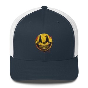 May include: A navy blue trucker hat with a white mesh back. The front of the hat features a yellow smiley face with a sinister grin and blood-like splatters. The smiley face has a cross over the eyes.