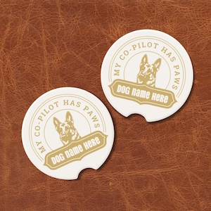 May include: Two white car coasters with a German Shepherd illustration and the text "MY CO-PILOT HAS PAWS" and "DOG NAME HERE" in gold. The coasters are round with a small notch on one side.