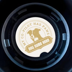 May include: A white and gold car coaster featuring a dog illustration and the text "MY CO-PILOT HAS PAWS" and "DOG NAME HERE". The coaster sits in a black car cup holder.