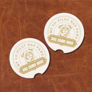 May include: Two cream-coloured car coasters featuring a pug illustration and the text "MY CO-PILOT HAS PAWS" and "DOG NAME HERE" in gold. The coasters are circular with a small notch.