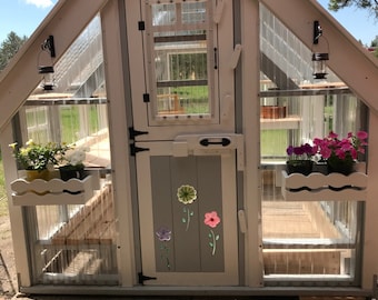 May include: A small, white wooden greenhouse with a gray door and a clear plastic roof. The door has a black handle and a black latch. There are two small windows on the door. There are flowers in pots on the shelves inside the greenhouse.