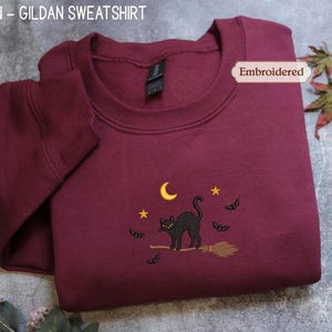 A burgundy sweatshirt with a black cat embroidered on it, featuring a broomstick and bats, is displayed on a gray surface alongside some leaves.