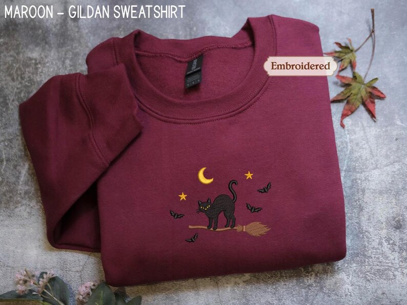 A burgundy sweatshirt with a black cat embroidered on it, featuring a broomstick and bats, is displayed on a gray surface alongside some leaves.