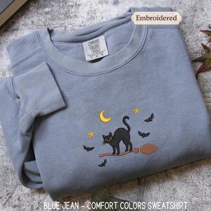 A gray sweatshirt with a black cat and bats on a broom, along with a label that says "Embroidered" and a tag that reads "Comfortable Colors Sweatshirt".