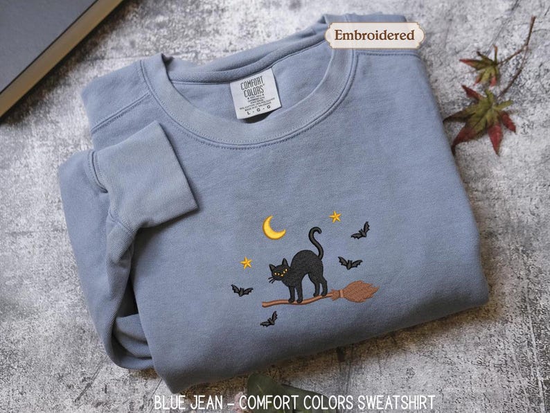 A gray sweatshirt with a black cat and bats on a broom, along with a label that says "Embroidered" and a tag that reads "Comfortable Colors Sweatshirt".
