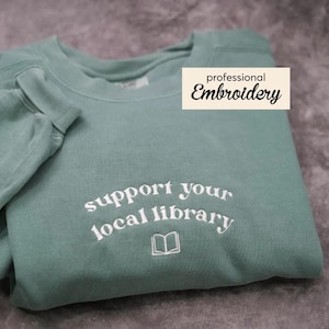 May include: A teal sweatshirt with white embroidered text saying "support your local library" and a book icon. The words "professional Embroidery" are printed on a white rectangle.