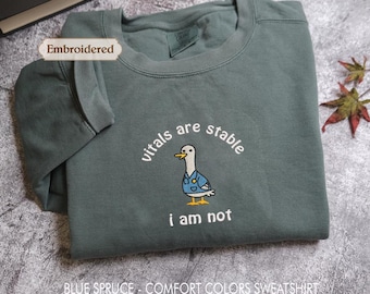 May include: A blue spruce crewneck sweatshirt featuring an embroidered graphic of a duck wearing a blue medical scrub top and a stethoscope. The text "vitals are stable i am not" is embroidered in white above and below the duck illustration.