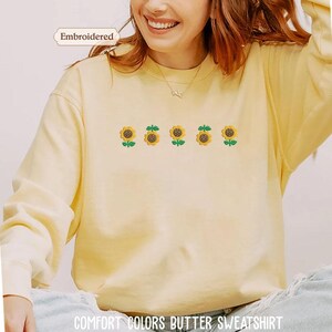 A woman wearing a yellow sweater with a row of five embroidered sunflowers on it.