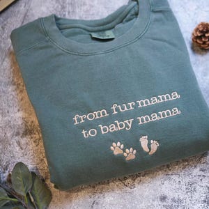 A gray sweatshirt with the text "from fur mama to baby mama" printed on it, along with paw prints, is displayed on a gray surface alongside some leaves and a pine cone.