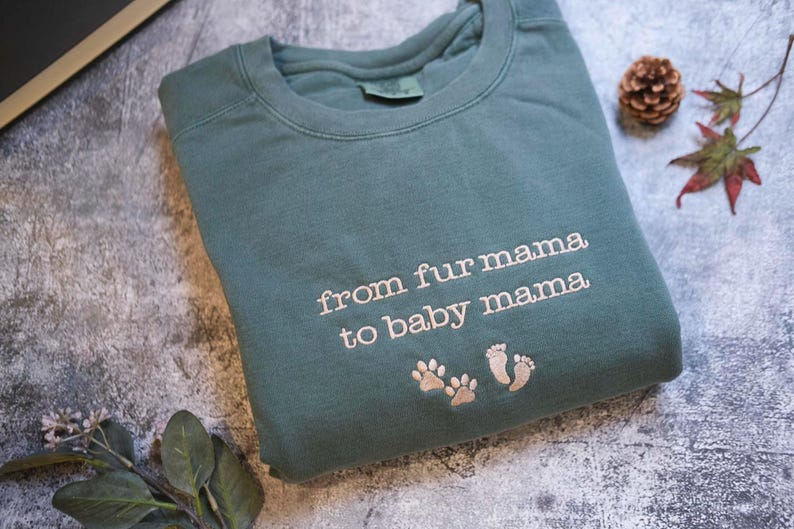 A gray sweatshirt with the text "from fur mama to baby mama" printed on it, along with paw prints, is displayed on a gray surface alongside some leaves and a pine cone.