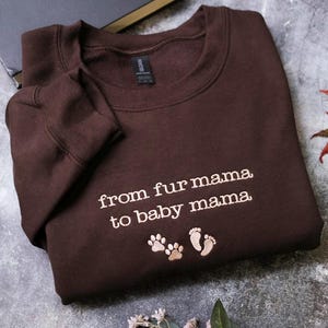 A brown sweatshirt with the text "from fur mama to baby mama" printed on it, along with paw prints, is laid out on a gray surface.