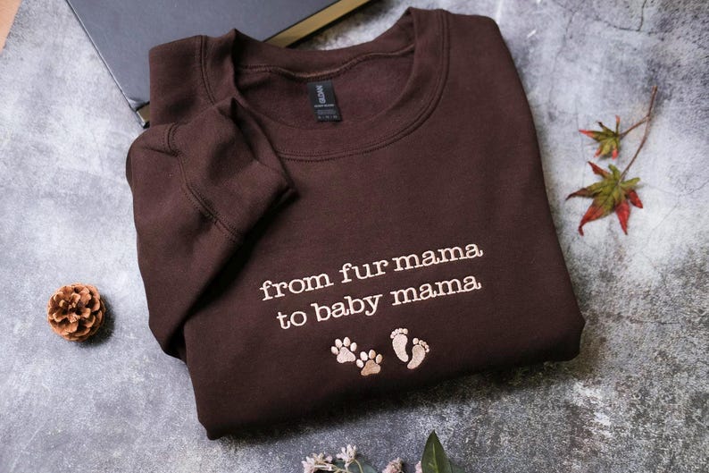 A brown sweatshirt with the text "from fur mama to baby mama" printed on it, along with paw prints, is laid out on a gray surface.