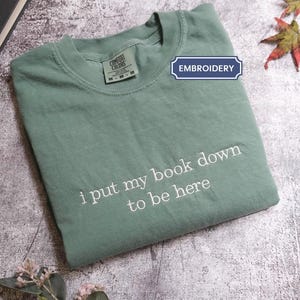 A green t-shirt with the text "I put my book down to be here" printed on it, along with a leaf and a book.