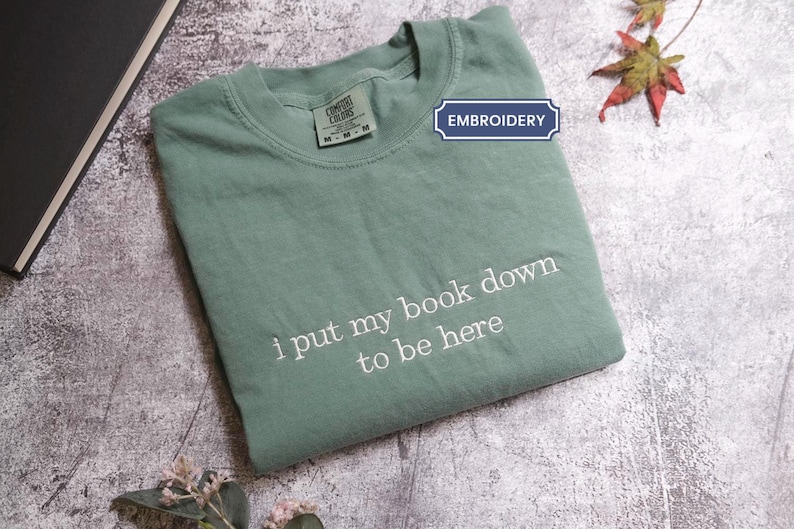 A green t-shirt with the text "I put my book down to be here" printed on it, along with a leaf and a book.