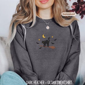 A woman wearing a gray sweatshirt with a black cat and bats embroidered on it, sitting on a couch.