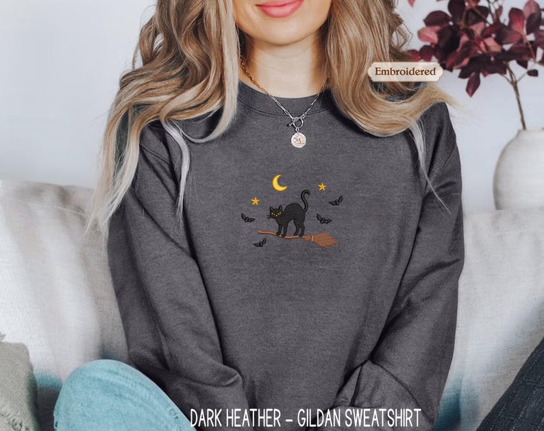 A woman wearing a gray sweatshirt with a black cat and bats embroidered on it, sitting on a couch.