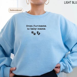 A person wearing a light blue sweatshirt with the text "from fur mama to baby mama" and paw prints embroidered on it.