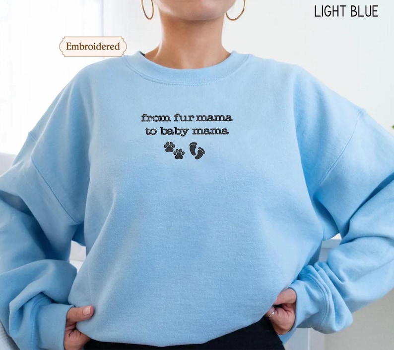 A person wearing a light blue sweatshirt with the text "from fur mama to baby mama" and paw prints embroidered on it.