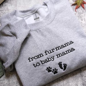 A gray sweatshirt with the text "from fur mama to baby mama" and paw prints is laid out on a surface.