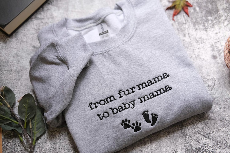 A gray sweatshirt with the text "from fur mama to baby mama" and paw prints is laid out on a surface.