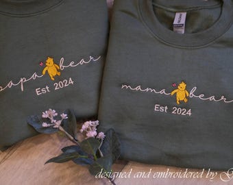 May include: Two olive green sweatshirts with embroidered designs. One says "papa bear" with a yellow bear and "Est 2024". The other says "mama bear" with a yellow bear and "Est 2024". A floral arrangement is in the foreground.