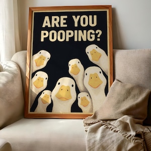 A framed poster with a humorous cartoon duck illustration and the text "ARE YOU POoping?" displayed on a couch.