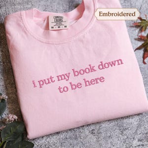 A pink t-shirt with the text "I put my book down to be here" embroidered on it, along with a label indicating it is "Embroidered".