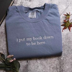 a blue t-shirt with the text "I put my book down to be here" printed on it, along with some leaves and a book.