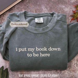 a gray sweatshirt with the text "I put my book down to be here" printed on it, along with a label that reads "Embroidered". The sweatshirt is laid out on a surface, and there are some leaves and a book visible in the background.
