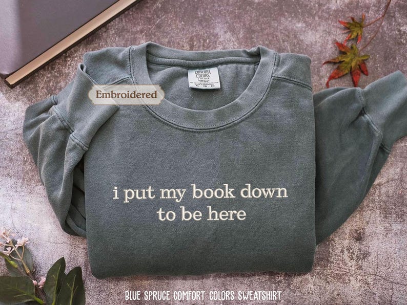 a gray sweatshirt with the text "I put my book down to be here" printed on it, along with a label that reads "Embroidered". The sweatshirt is laid out on a surface, and there are some leaves and a book visible in the background.