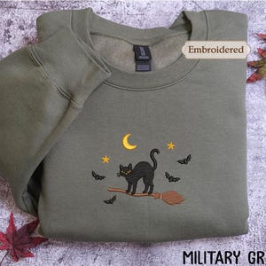 A military green sweatshirt with an embroidered design of a black cat on a broom, surrounded by bats and a crescent moon, with autumn leaves in the background.