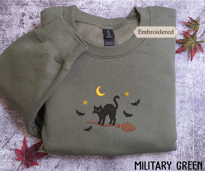 A military green sweatshirt with an embroidered design of a black cat on a broom, surrounded by bats and a crescent moon, with autumn leaves in the background.