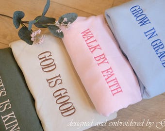 May include: Four folded sweatshirts in olive, beige, light pink, and light blue. The olive sweatshirt reads JESUS IS KING, the beige reads GOD IS GOOD, the pink reads WALK BY FAITH, and the light blue reads GROW IN GRACE in embroidery.