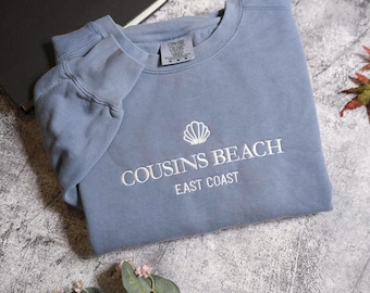 Può includere: Una felpa blu con la scritta bianca ricamata "COUSINS BEACH EAST COAST" e un motivo a conchiglia. Il capo è piegato su una superficie strutturata, con un libro e foglie autunnali sullo sfondo.