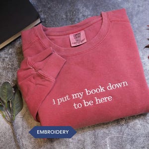 a red long-sleeved shirt with the text "I put my book down to be here" printed on it, along with a black book and some leaves.